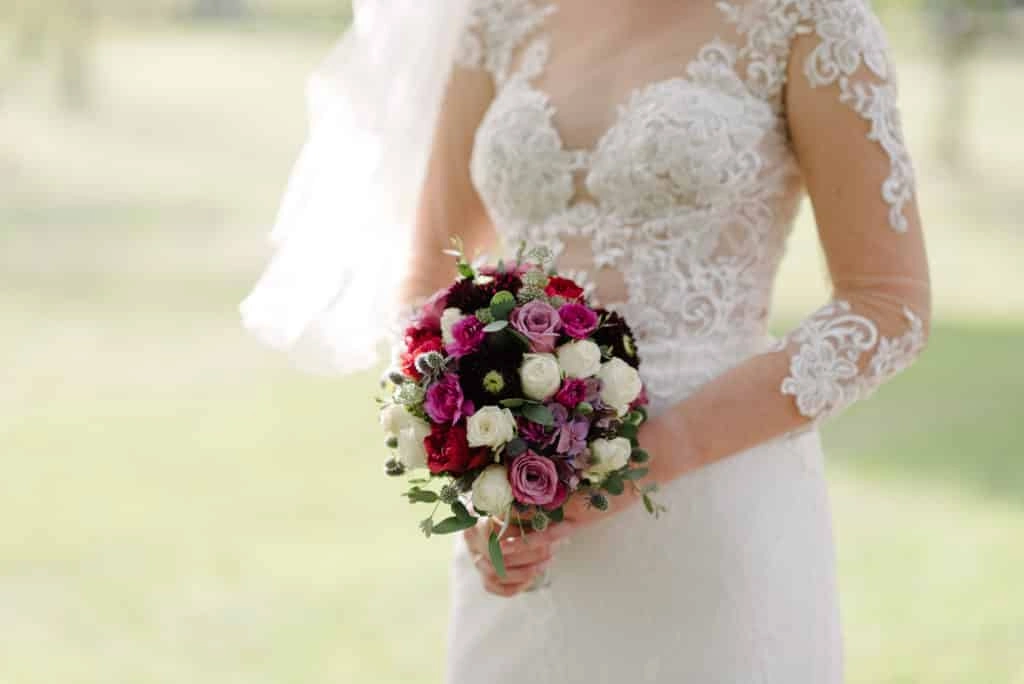 Brautstrauß mit Rosen vor einem weißen Spitzenkleid.
