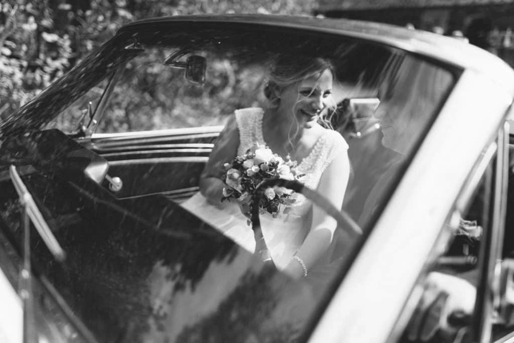 Die Braut sitzt mit ihrem Brautstrauß im Oldtimer; Schwarzweißaufnahme durch die Scheibe.