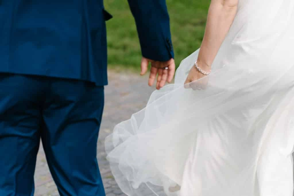 Braut und Bräutigam halten beim Gehen die Hände; das Brautkleid weht im Wind.