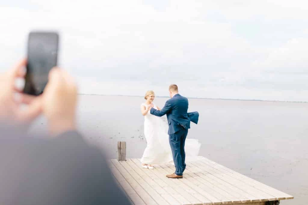 Ein Gast fotografiert das Brautpaar auf einem Holzsteg am Meer.