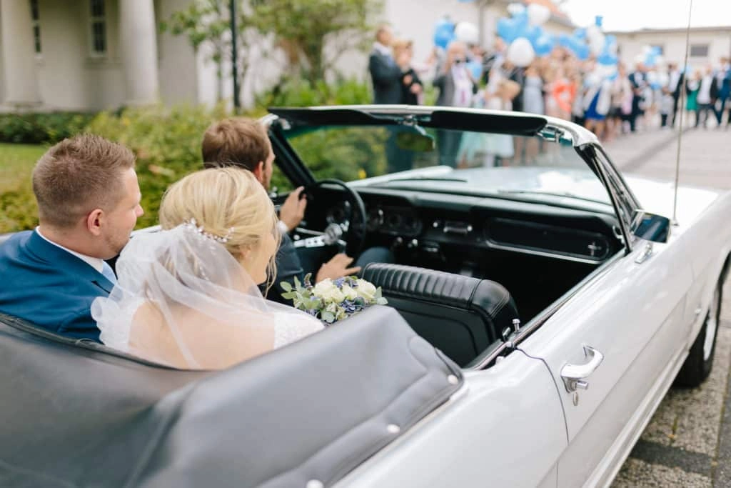 Das Brautpaar sitzt im offenen Hochzeitsauto, während die Gäste dahinter warten.