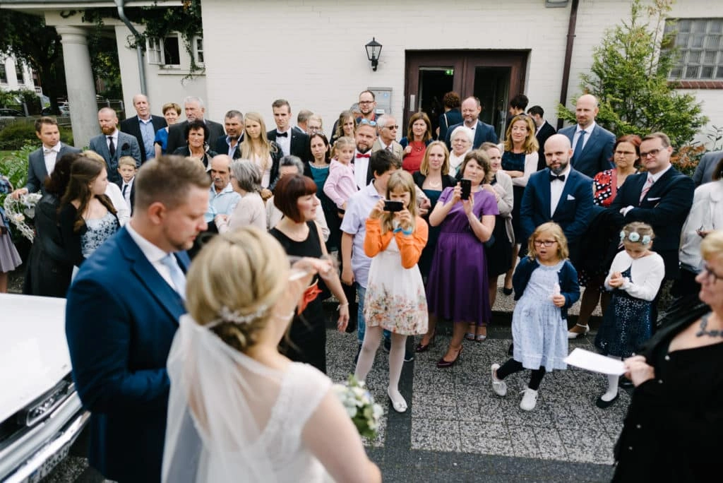 Hochzeitsgäste begrüßen das Brautpaar draußen vor dem Traugebäude.
