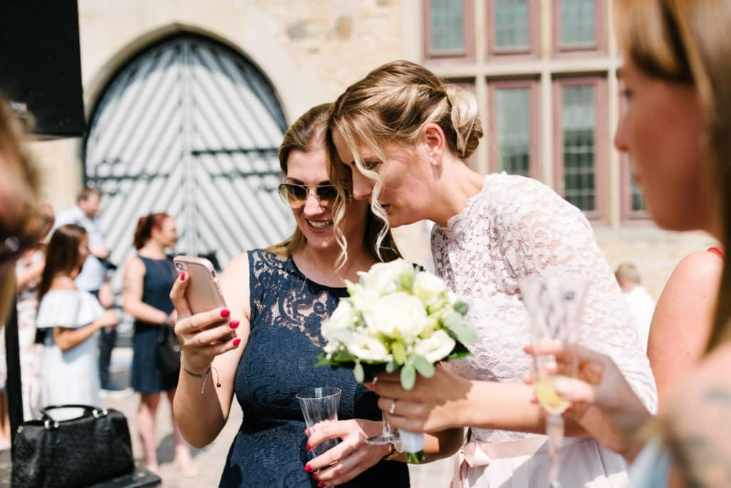 Braut betrachtet mit einem Gast ein Smartphone.