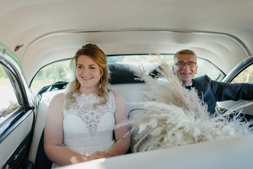 Die Braut sitzt mit Blumenstrauß und Begleitung im Oldtimer.