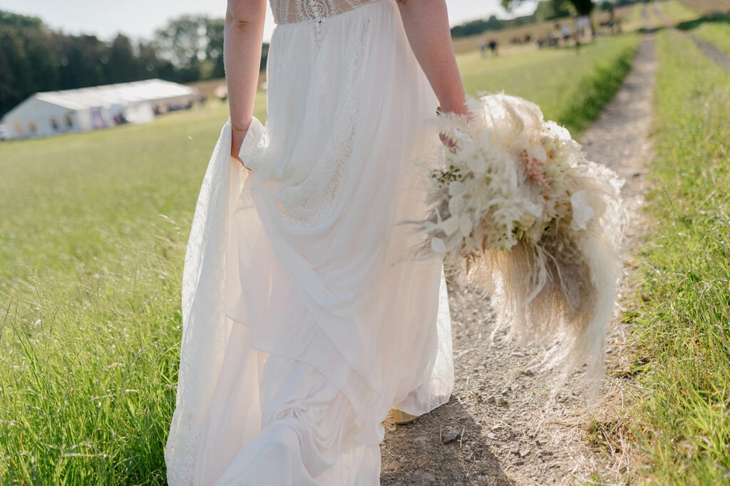 Detail von Brautkleid und Trockenblumenstrauß auf einem Feldweg.