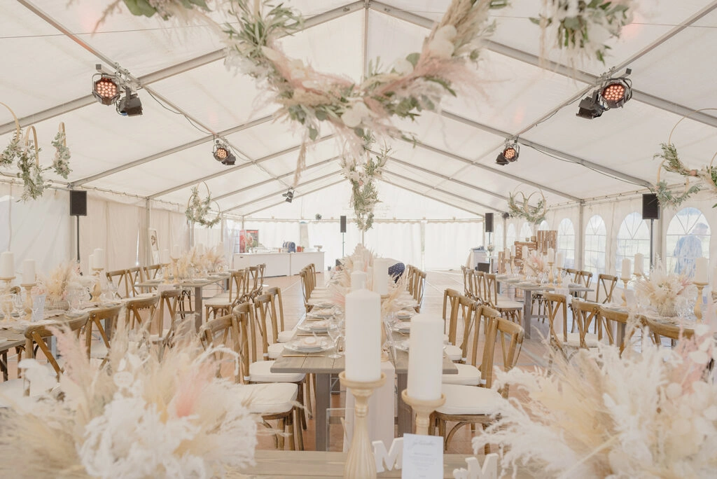 Lange Hochzeitstafeln im hellen Festzelt mit hängenden grünen Kränzen.