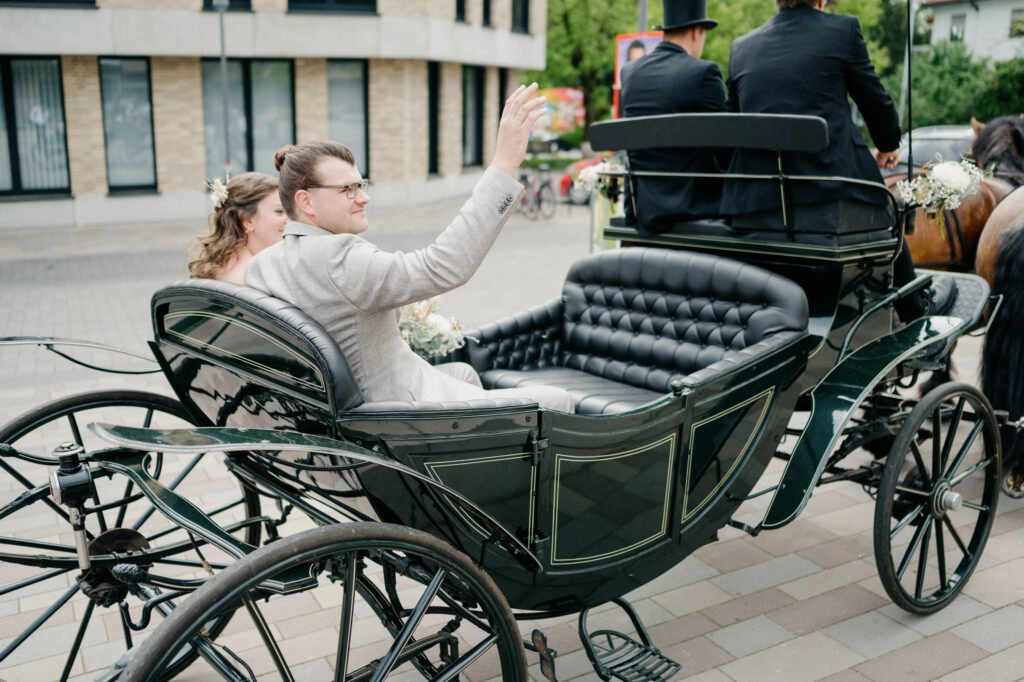 Brautpaar winkt aus einer offenen Pferdekutsche.