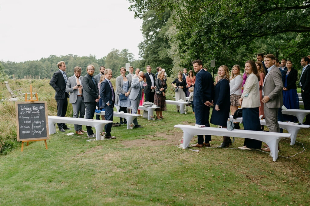 Hochzeitsgäste stehen zwischen den Bänken auf der Wiese.
