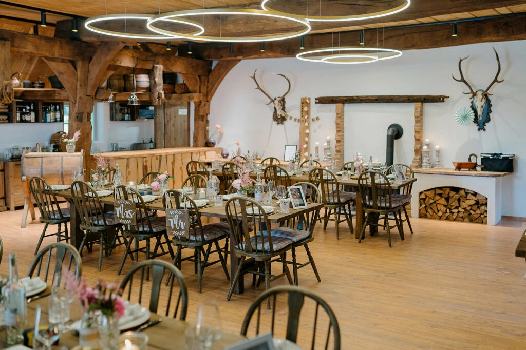 Festsaal mit Holztischen, Blumen und runden Deckenleuchten.