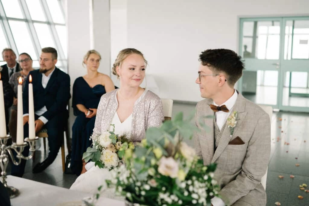 Brautpaar sitzt mit Blumenstrauß vor den Hochzeitsgästen.