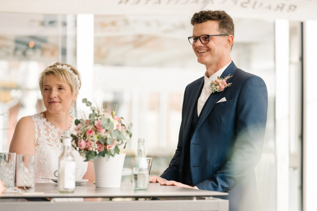 Brautpaar steht mit Blumenstrauß am gedeckten Tisch.