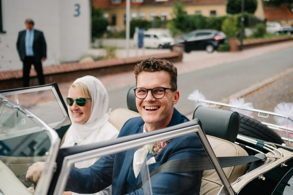 Brautpaar sitzt lachend im offenen Oldtimer.