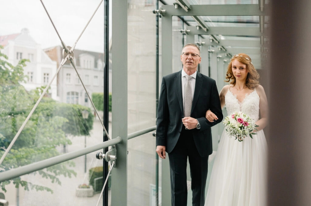 Braut geht mit Begleitung durch einen Glasgang.