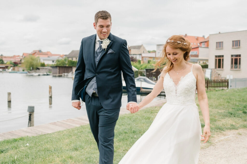Brautpaar spaziert Hand in Hand am Wasser.
