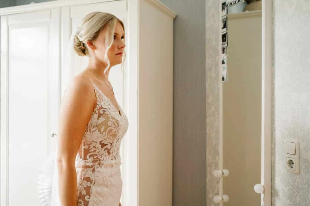 Braut im Spitzenkleid vor einem hellen Fenster.