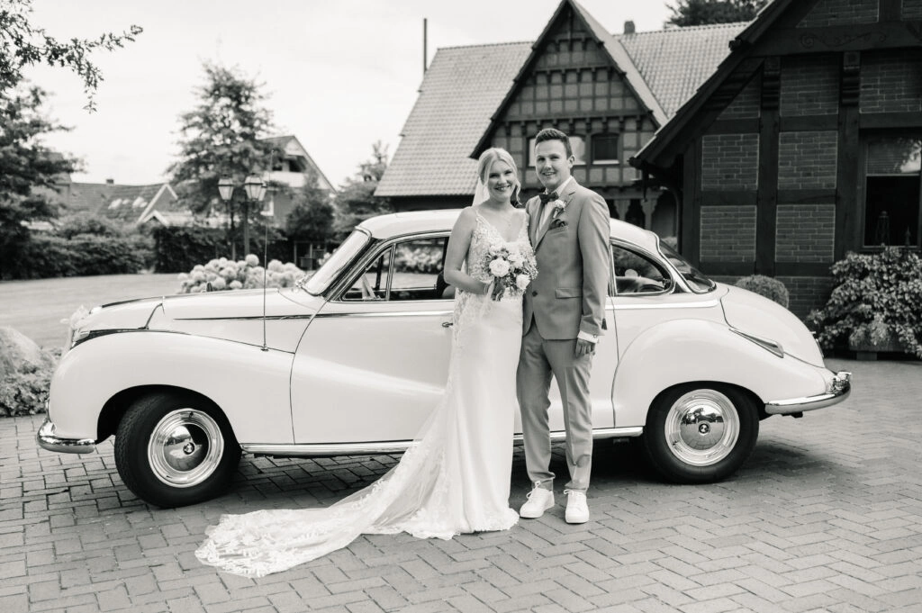 Brautpaar steht vor einem weißen Oldtimer und Fachwerkhäusern.