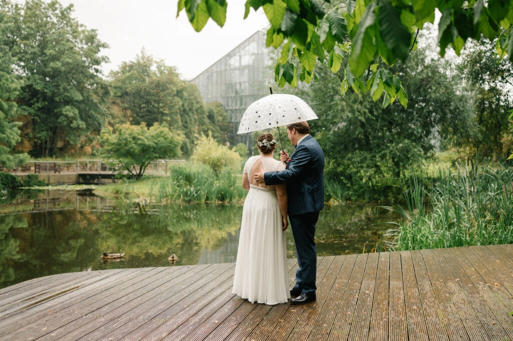 Brautpaar steht mit Regenschirm auf einem Holzsteg am Teich.
