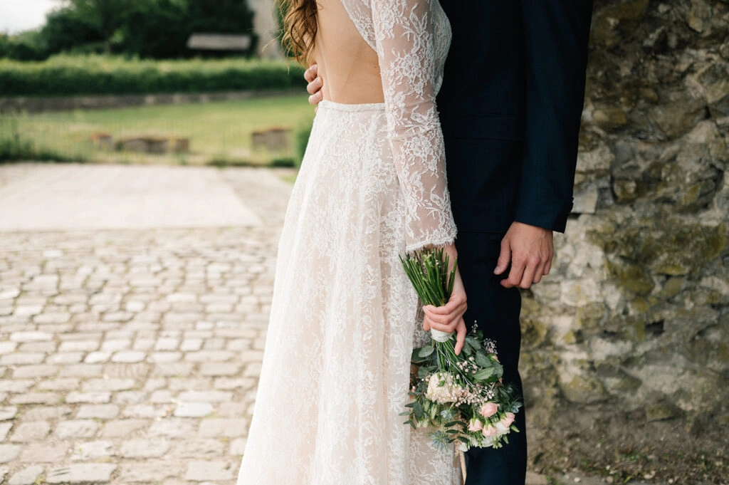 Detail von Spitzenkleid, Brautstrauß und dunklem Anzug.