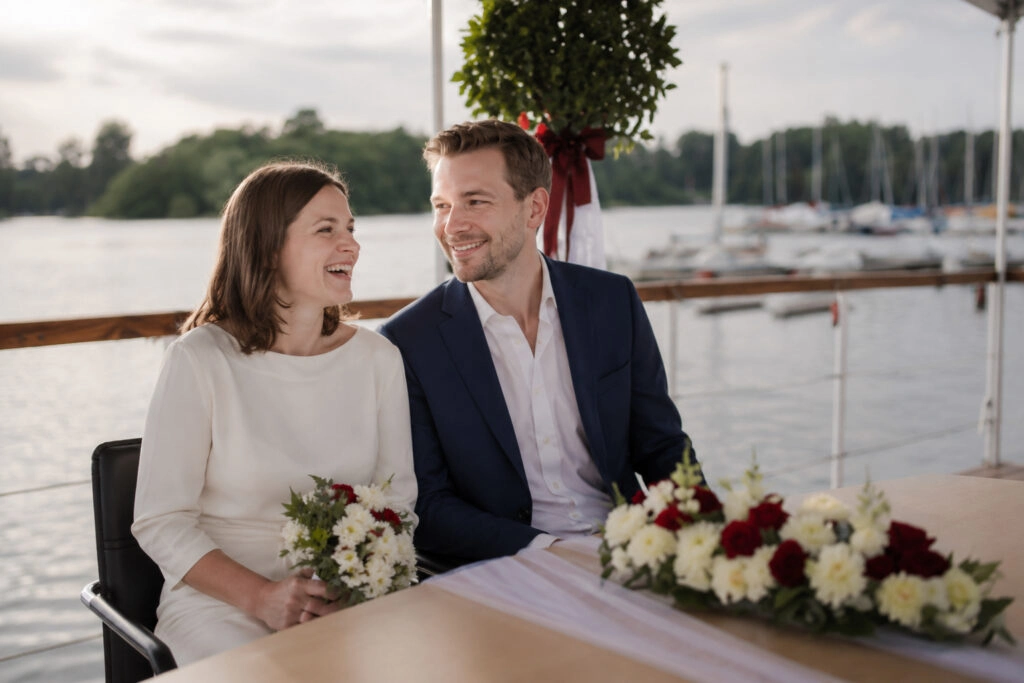 Braut und Bräutigam lächeln sich am mit Blumen geschmückten Trautisch an.