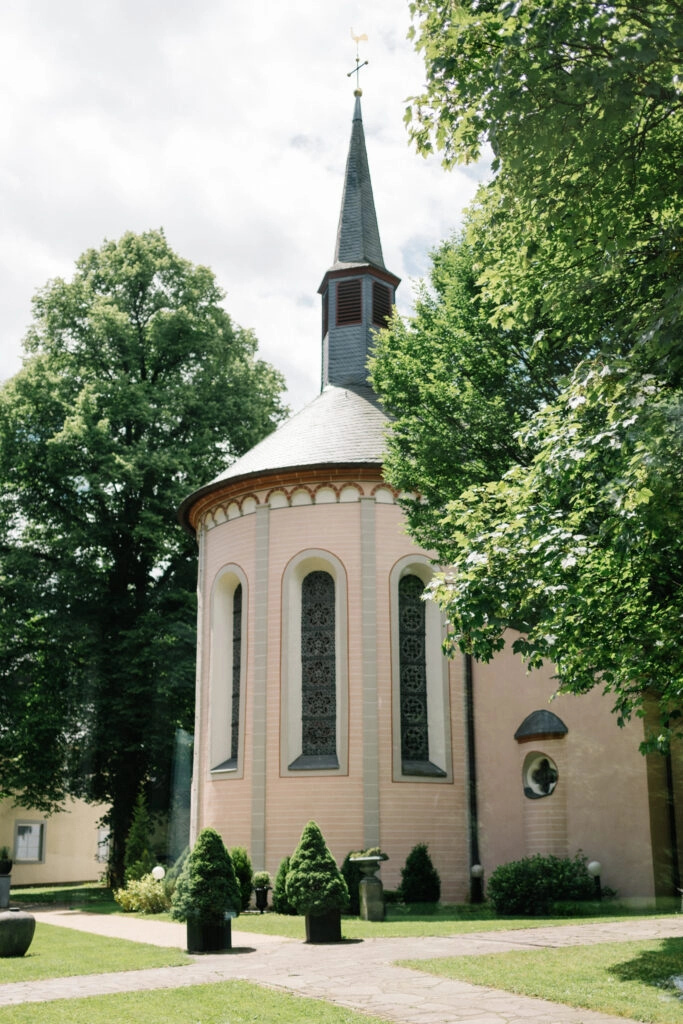 Kleine Kirche mit schmalem Turm zwischen Bäumen.