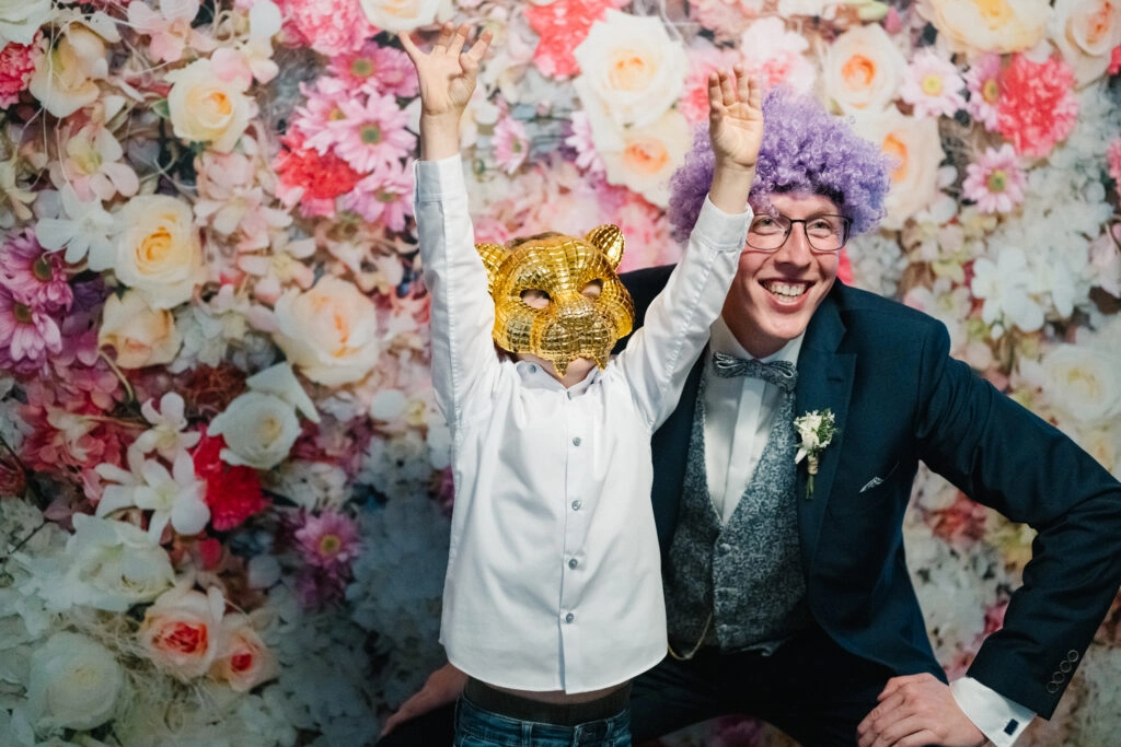 Bräutigam und Gast posieren mit Verkleidung vor einer Blumenwand.