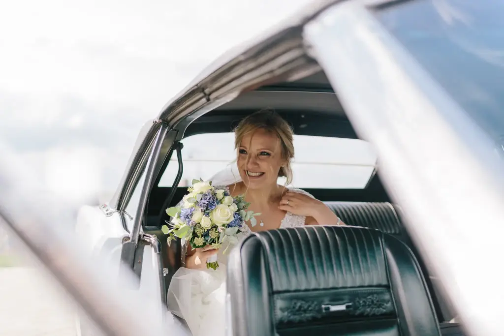 Eine lächelnde Braut sitzt mit ihrem Blumenstrauß auf dem Rücksitz eines Hochzeitsautos.