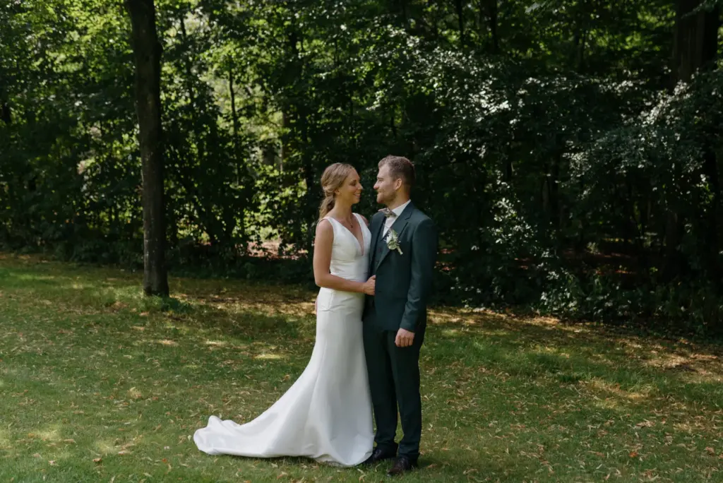 Ein Brautpaar steht im grünen Park; die lange Schleppe des Brautkleides liegt auf dem Rasen.