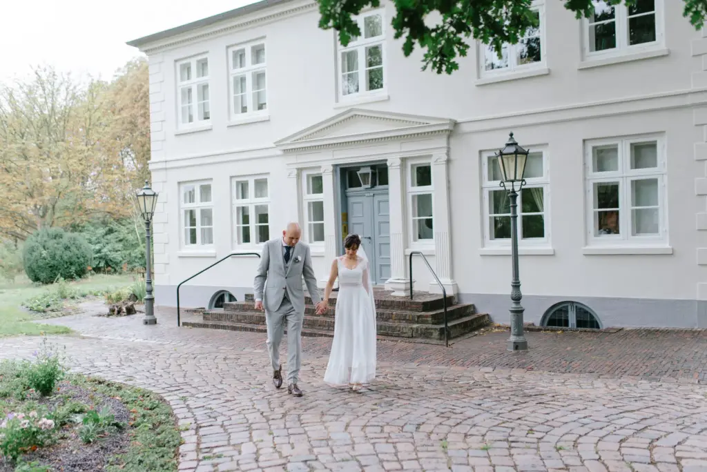 Ein Brautpaar geht vor einem weißen Herrenhaus über den gepflasterten Vorplatz.