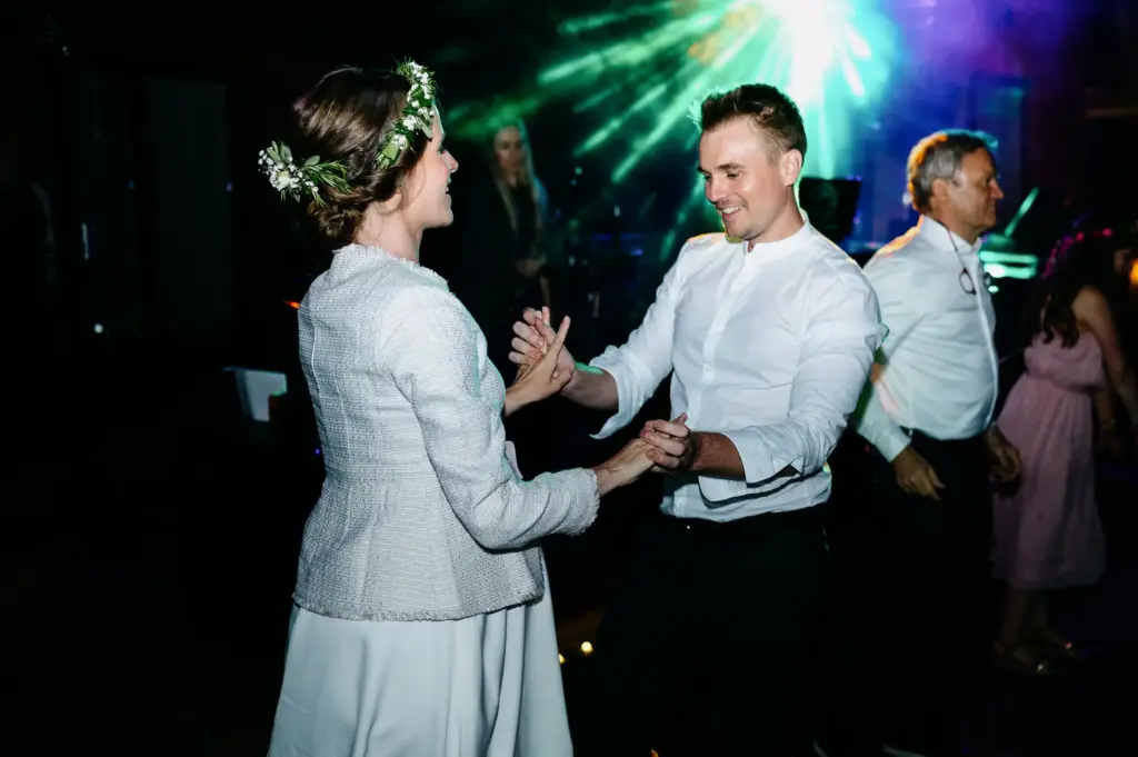 Ein Brautpaar tanzt lachend vor Gästen im grünen und blauen Partylicht.